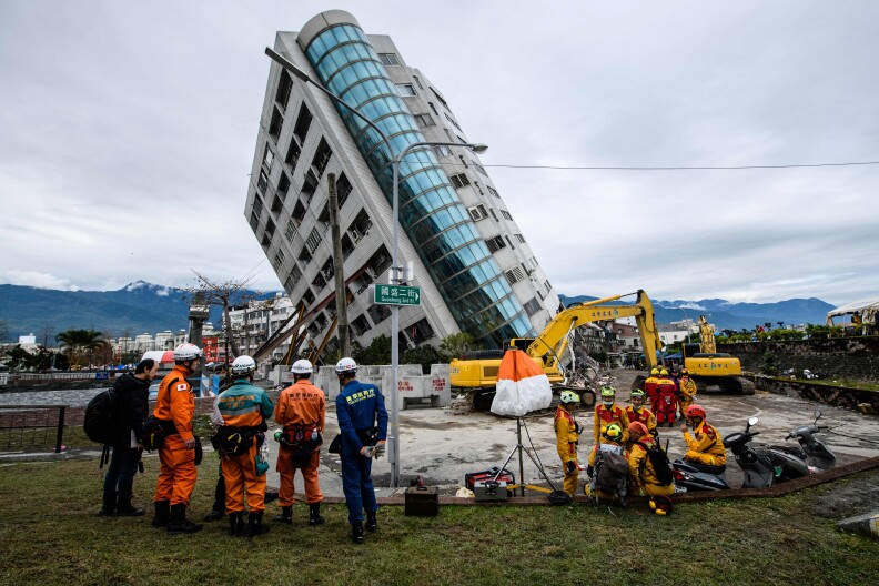 Rescue workers in full uniforms of orange and dark blue stand and observe a building leaning heavily to the left on the brink of collapse.