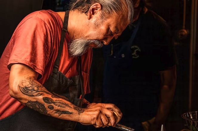 A man with a medium-dark skin tone, long gray hair in a ponytail, and a long gray goatee leans over his work at the counter, his tattooed arms visible as he prepares a dish.