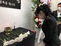 A woman dressed in black bows her head in front of an altar with more than half a dozen white chrysanthemums, four candles, and incense burning in an urn.