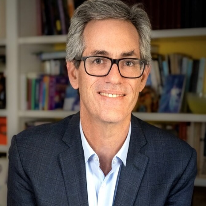 A man with light-tons skin and salt-and-pepper hair poses in front of a bookcase.