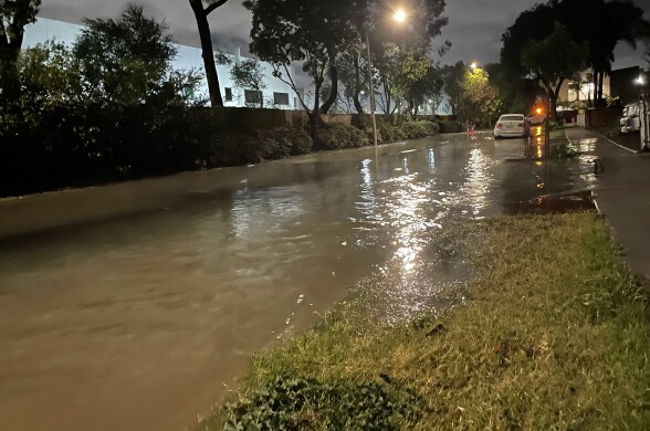 Sewage water flooding a city/neighborhood street by a sidewalk. The water is brown and murky. There is a car parked down the road.