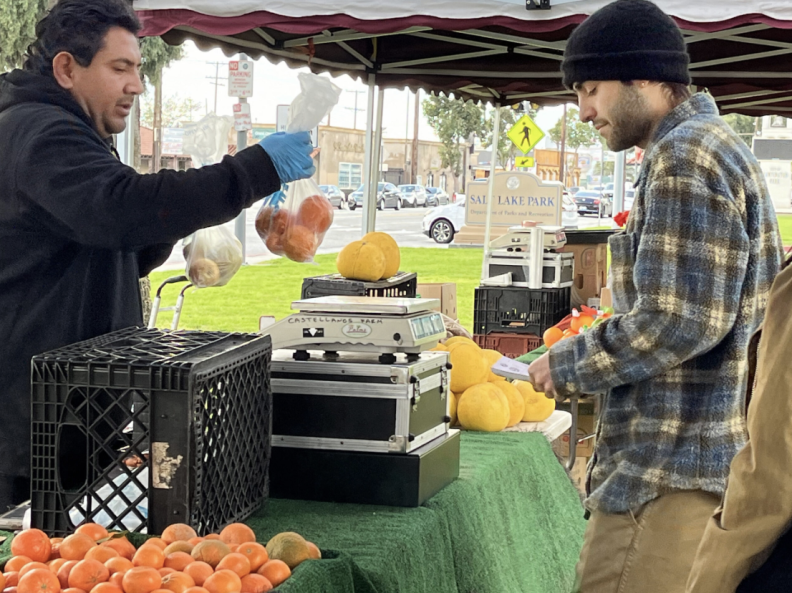 At a farmers market, a man dressed in a black hooded sweatshirt on the left is holding up a clear bag of oranges in his right gloved hand. In front of the man is another person with light skin tone dressed in a thick long-sleeved shirt and a beanie cap. They are both standing at a table that has a milk crate and a small scale that sits atop a couple of cases.