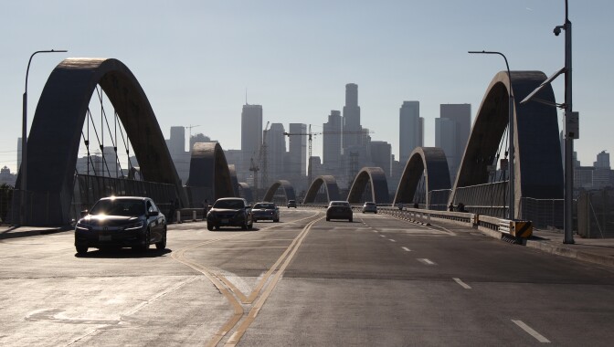 6 cars drive across a bridge with arches on both sides of it. In the distance is the Downtown Los Angeles skyline with high rises and office buildings.