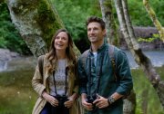 A man and woman stand together, each wearing backpacks and holding binoculars down at their waists. Behind them is a mossy tree trunk, grass and trees.