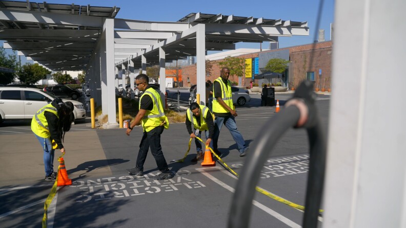 Four people wearing neon safety vests do work in a parking lot on a sunny day. The parking spaces read "electric vehicle chargin" and a portion of an electric vehicle charger can be seen in the foreground. 