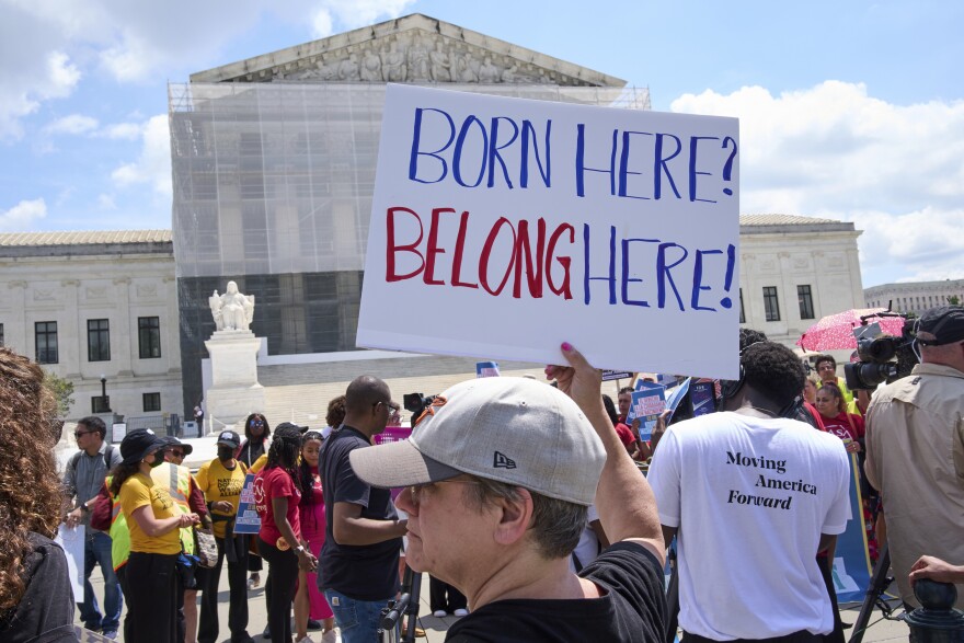 A person holds a sign that reads "Born here? Belong here!" around other people, some holding signs, outside the Supreme Court, which is under construction.