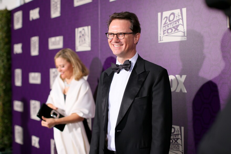 Peter Rice, wearing a tuxedo, stands in front of a purple 20th Century Fox step-and-repeat background. A blonde woman in white is behind him, to his right.