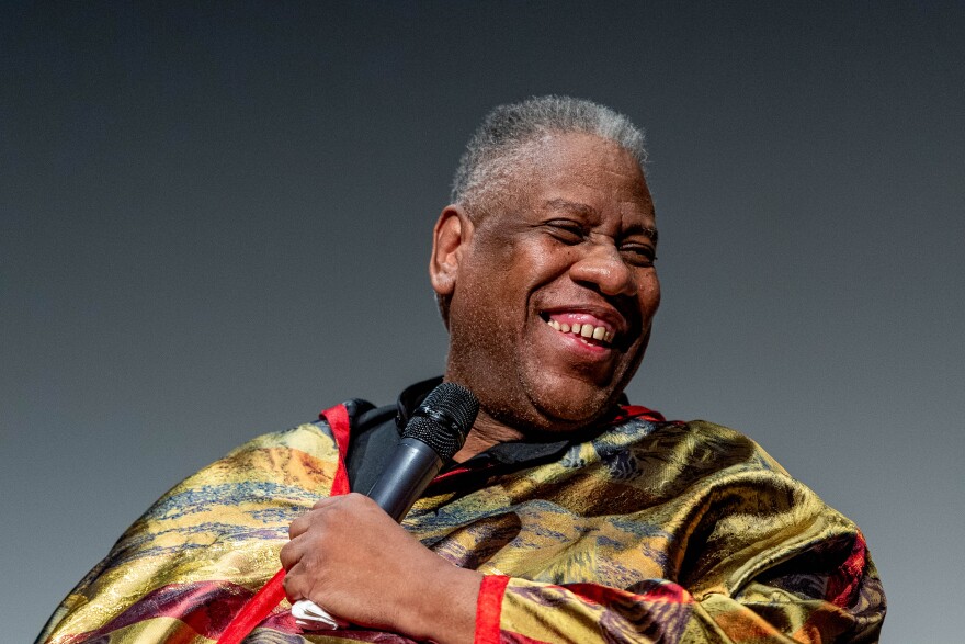 A close-up of Andre Leon Talley with a vibrant outfit holding a microphone. He has a wide smile and is looking off to the side.