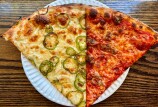 Two large pizza slices side by side on a white paper plate — one topped with jalapeño rounds and melted cheese, the other with tomato sauce, melted cheese, and bits of sausage — sitting on a wooden table.