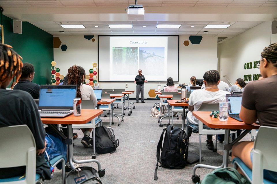 Students sit in a crowded classroom while a professor stands and lectures the class.