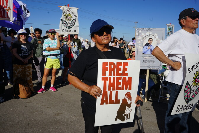 A diverse group of people protest outside. The most prominent sign in picture reads: Free Them All.