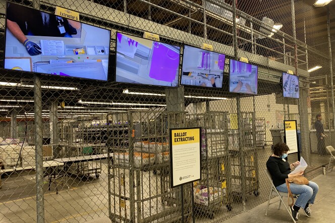 Monitors are attached to a chain-link fence inside a warehouse. The monitors show people's handling ballots.