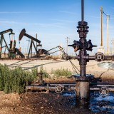 Several oil derricks and a pipeline surrounded by a pool of oil are shown in a dusty field