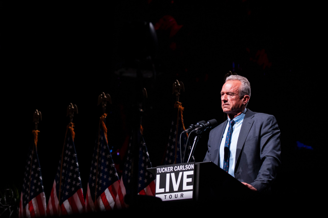An old man stands behind a podium that says "Tucker Carlson Live Tour". There are American flags in the background