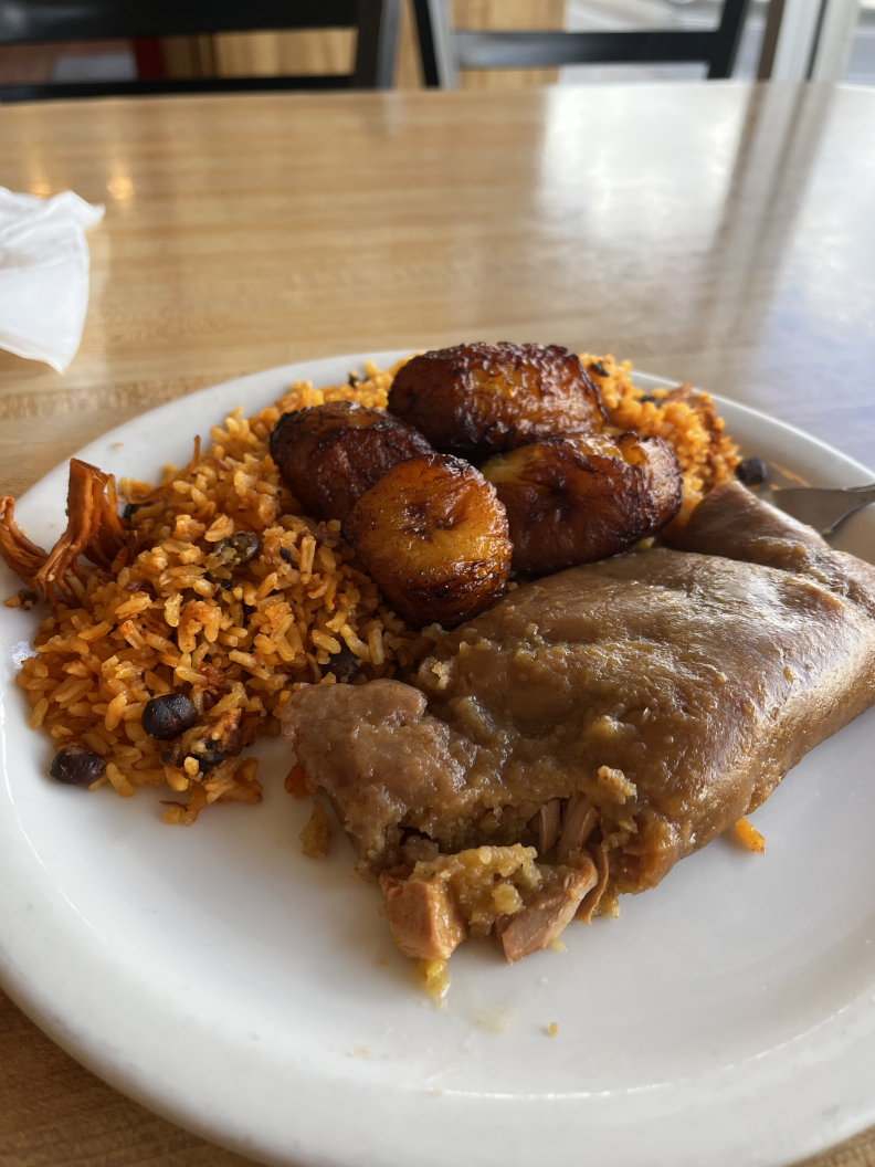 A plate sits on a table top, containing a pastele with a serving of fried plantains over a bed of orange rice
