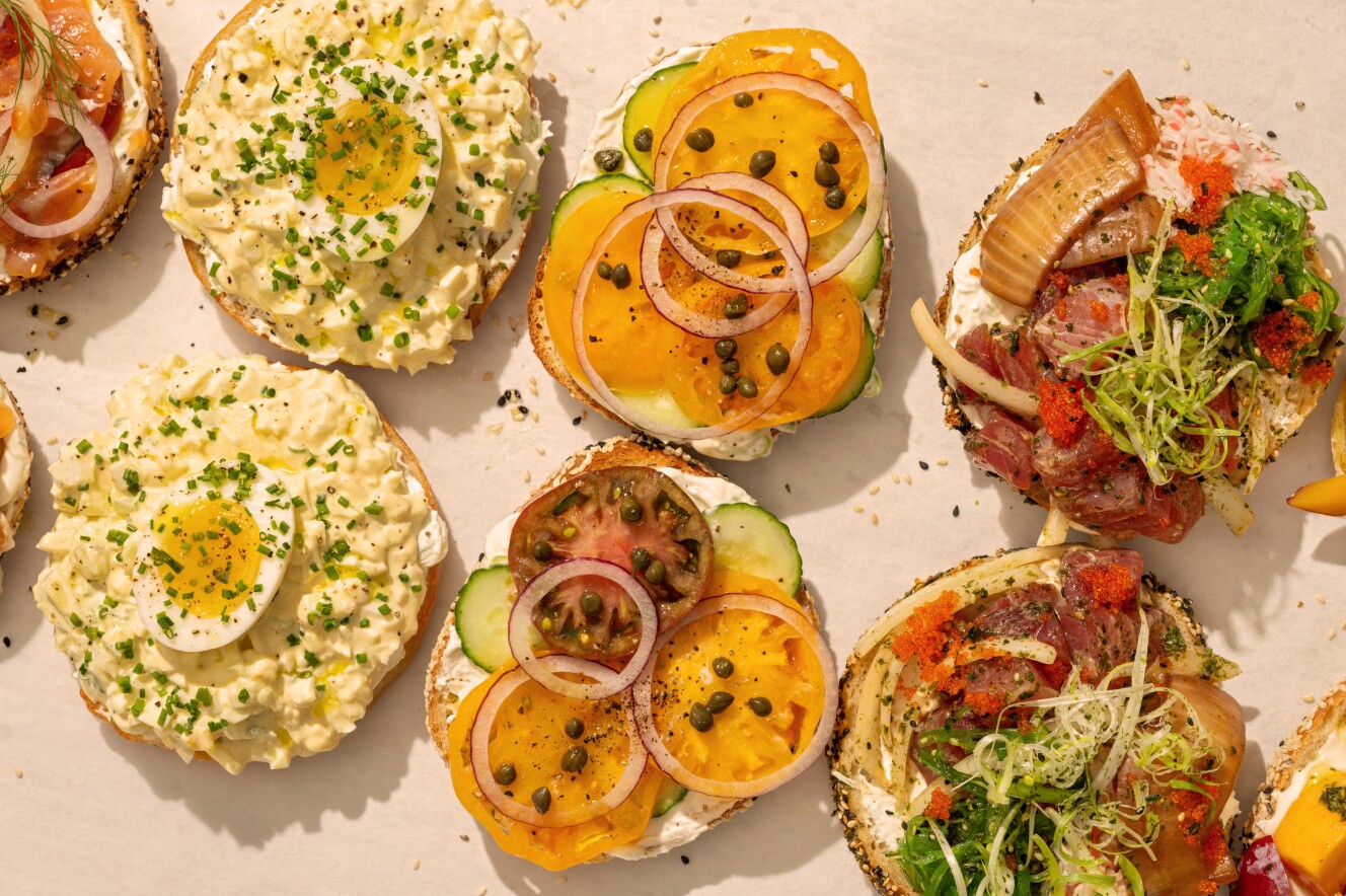Overhead view of several open-face bagels topped with different spreads, including egg salad with half a boiled egg, heirloom tomatoes with cucumbers and capers, and a poke-style mix with seaweed salad, smoked fish, onions, and roe, all arranged on parchment.