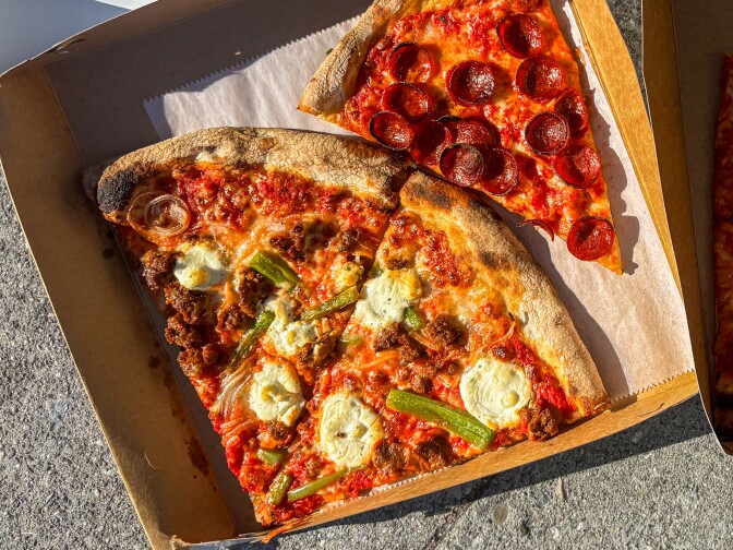 Two slices of pizza in a takeout box: one topped with pepperoni, and the other with sausage, onions, dollops of ricotta, and green peppers, both on thin, charred crusts.