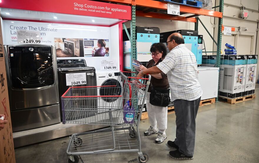 A man and woman pushing a shopping cart inspect washers and dryers inside a big box store.