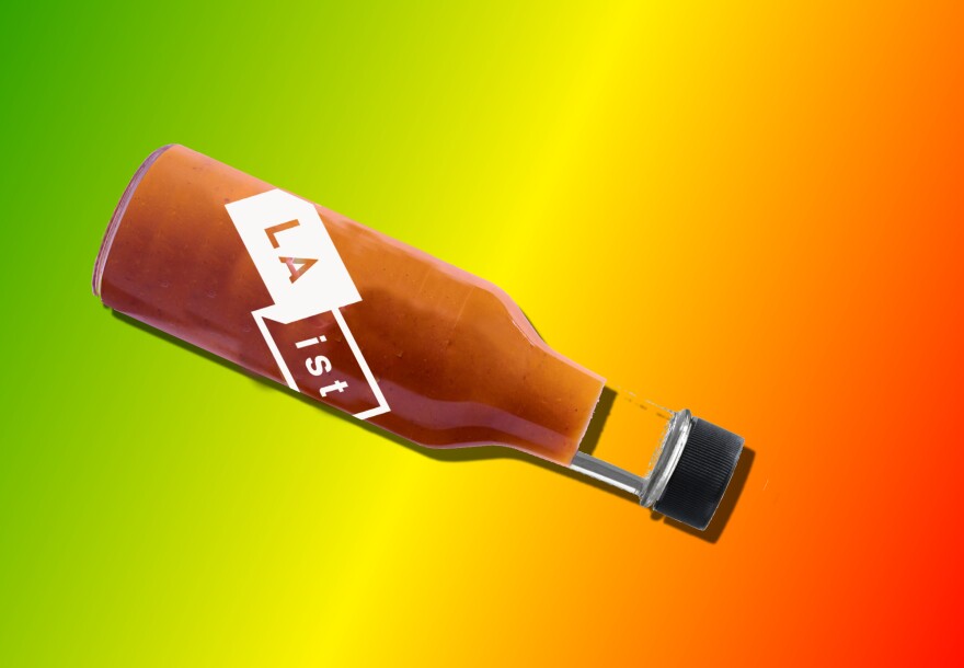 A bottle of red hot sauce with the LAist logo, tipped on its side, against a green, yellow, orange and red background