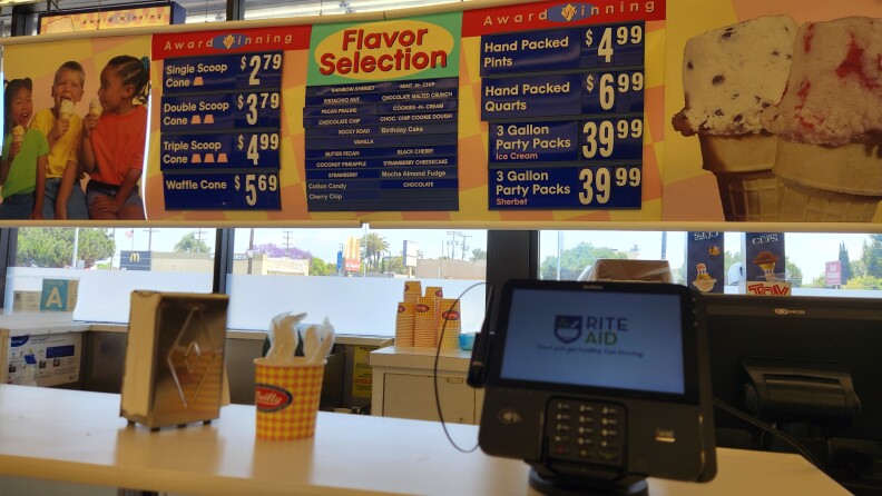 A Thrifty icecream counter in a Rite Aid. At the back is the ice cream menu, showing a single scoop cone for $2.79, and the prices increasing as the sizes get bigger. On the counter there is an electric terminal, a cup holding plastic spoons and a napkin holder.