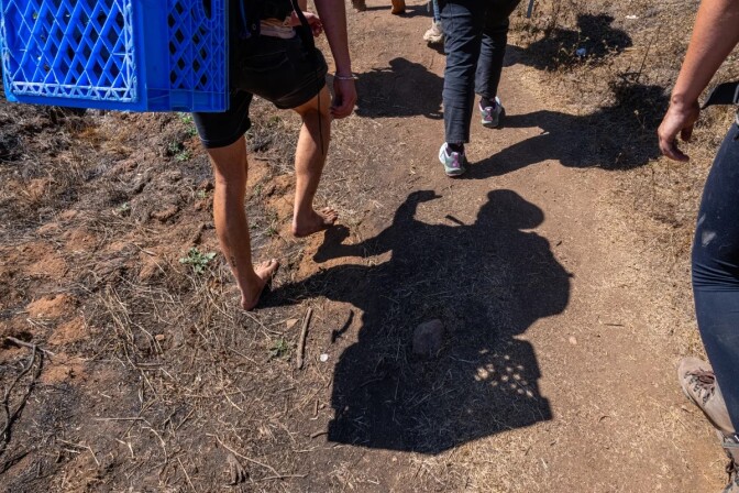An image depicts the feet of migrants walking along a dirt path with some holding objects 