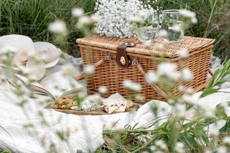 A wicker basket in a field on a white blanket and flowers.