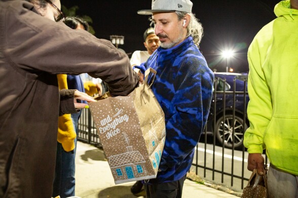 It's night. A man with a gray beard and a gray cap hold a paper grocery bag. A woman with a brown jacket reaches in.
