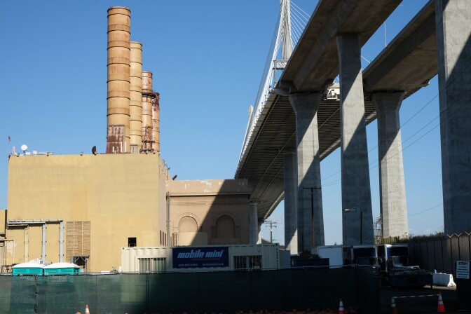A large power generating station next to a huge concrete bridge under a sunny blue sky. 