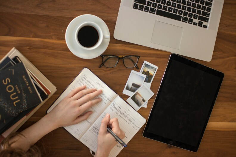 A woman writing in a journal with a tablet and a laptop nearby as well as a pair of glasses, a cup of coffee, and polaroid pictures spread around.