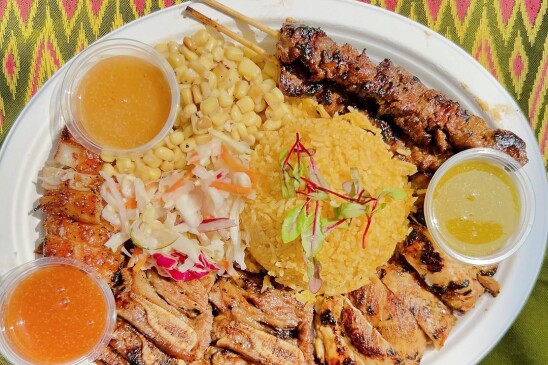 A plate of barbecued meat, including two skewer, with rice coleslaw and three sauces, displayed on a white plate sitting atop a greek cloth with pink and yellow designs. 