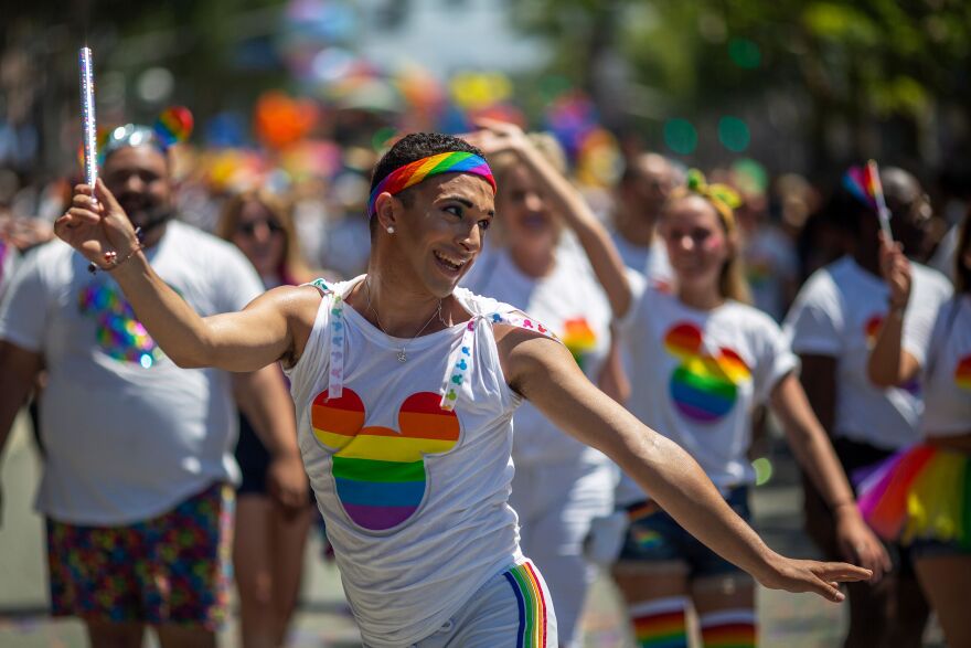 A man in a rainbow headband and a rainbow Mickey Mouse T-shirt holds up a baton while marching in a parade. Other men and women are behind him in rainbow Mickey gear, blurred in the background.