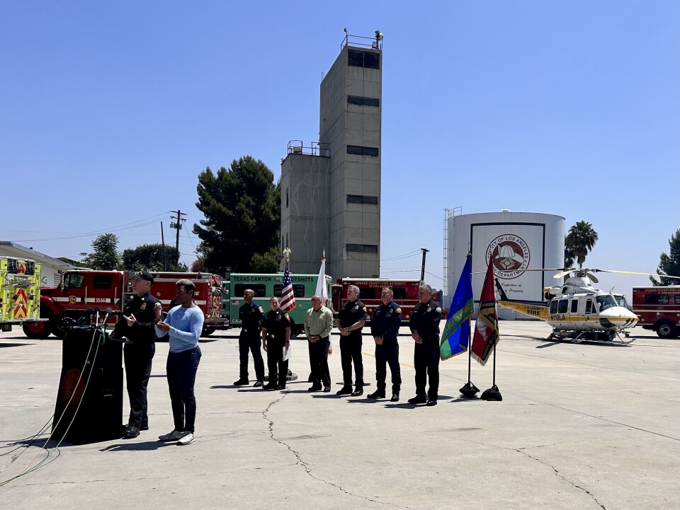 A man in black hat and black official uniform is speaking into microphones on a podium towards the left of the image, with a man wearing a blue sweater and jeans doing American Sign Language to the right of him. Behind them both are five men and one woman wearing similar black uniforms, with their hands clasped in front of them, standing in front of several different flags. A helicopter and several different fire engines are displayed behind them.