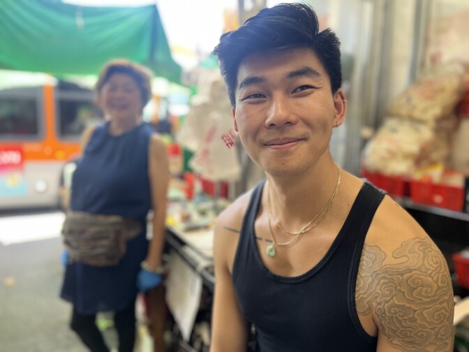 A younger Asian man in a black tank top sits smiling, showing a cloud-pattern tattoo on his shoulder. Behind him,  an older Asian woman in a blue tank top stands under a green tarp with a Metro bus passing on the street outside.