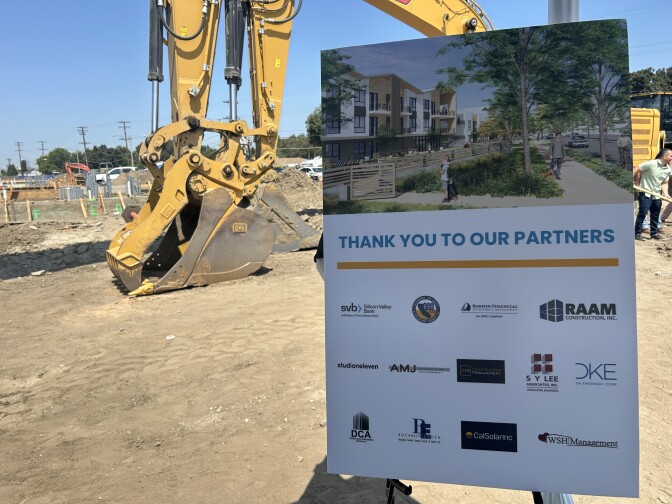 A sign in the foreground shows a rendering if an apartment complex with trees and grass out front and the words "Thank you to our partners" with company names and logos below. In the background are earth movers on a dirt lot. 