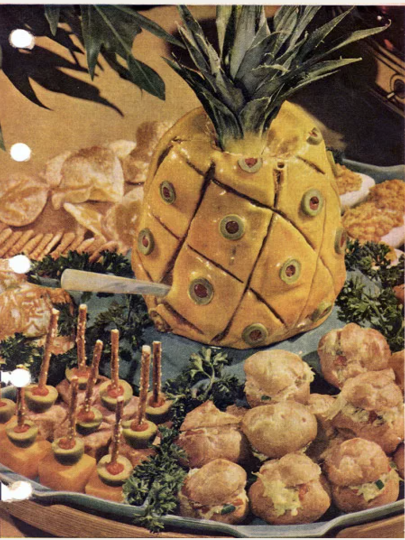 A pineapple-looking sculpture that's yellow and covered with green olives stuffed with red pimento. Surrounding the pineapple are other various appetizers