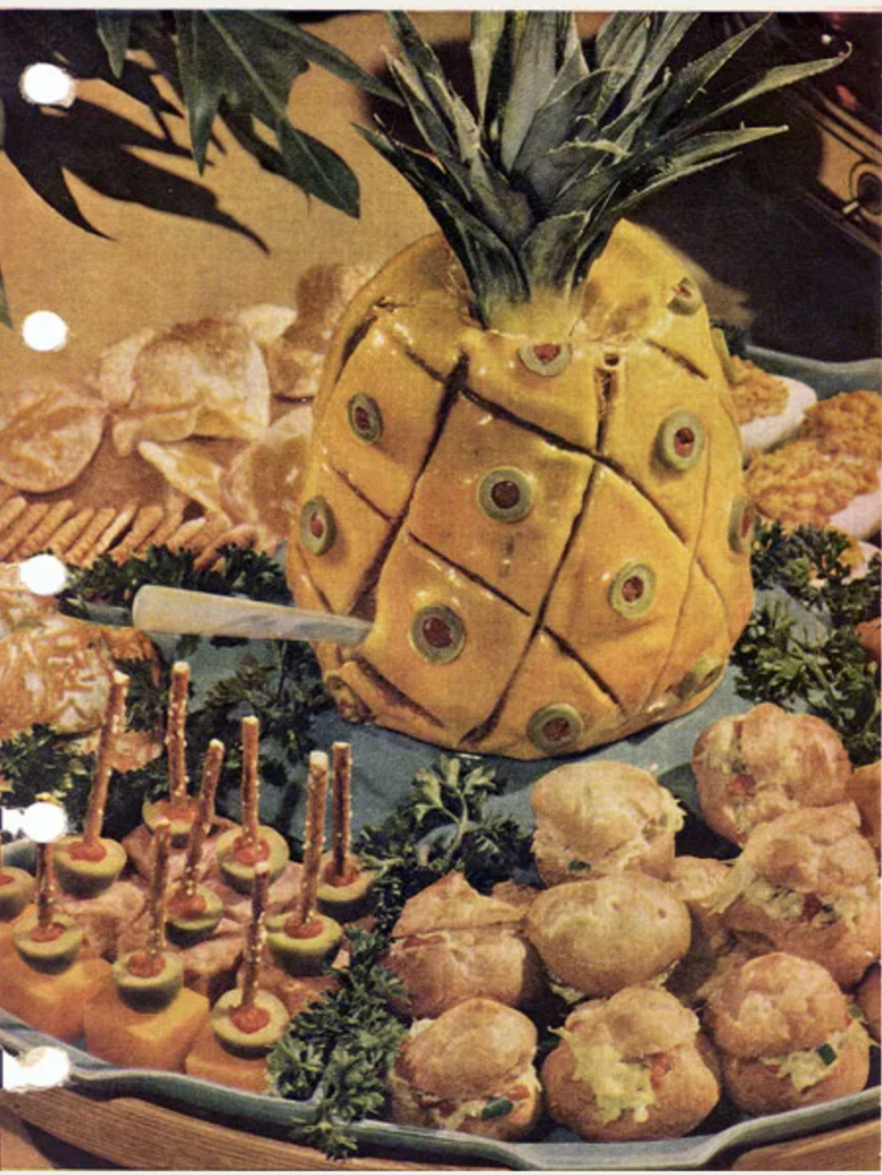 A pineapple-looking sculpture that's yellow and covered with green olives stuffed with red pimento. Surrounding the pineapple are other various appetizers 