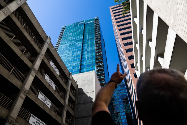 A man in the bottom right corner of the frame is slightly out of focus as he points up towards a blue skyscraper with a facade of windows.