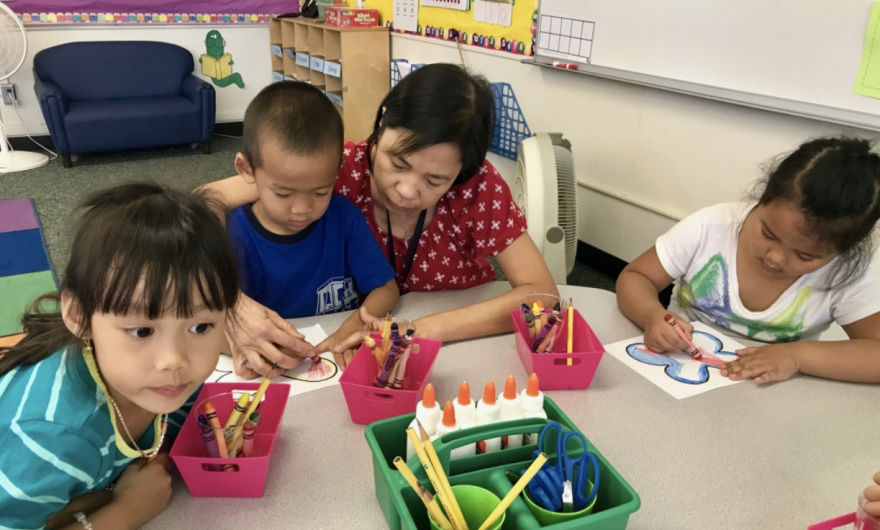 An Asian woman in a red short-sleeved, polka-dot shirt helps three kindergarten students who are coloring with crayons.