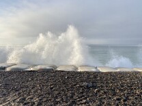 White spray from waves flies into the air above a line of large rectangular-shaped white bags. In the foreground there's a strip of gravel. 