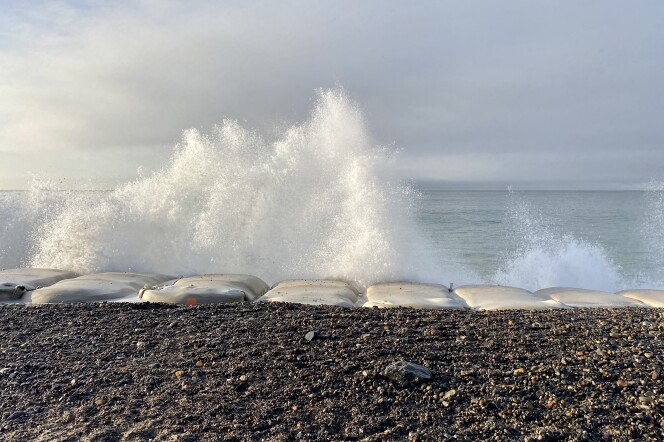 White spray from waves flies into the air above a line of large rectangular-shaped white bags. In the foreground there's a strip of gravel. 
