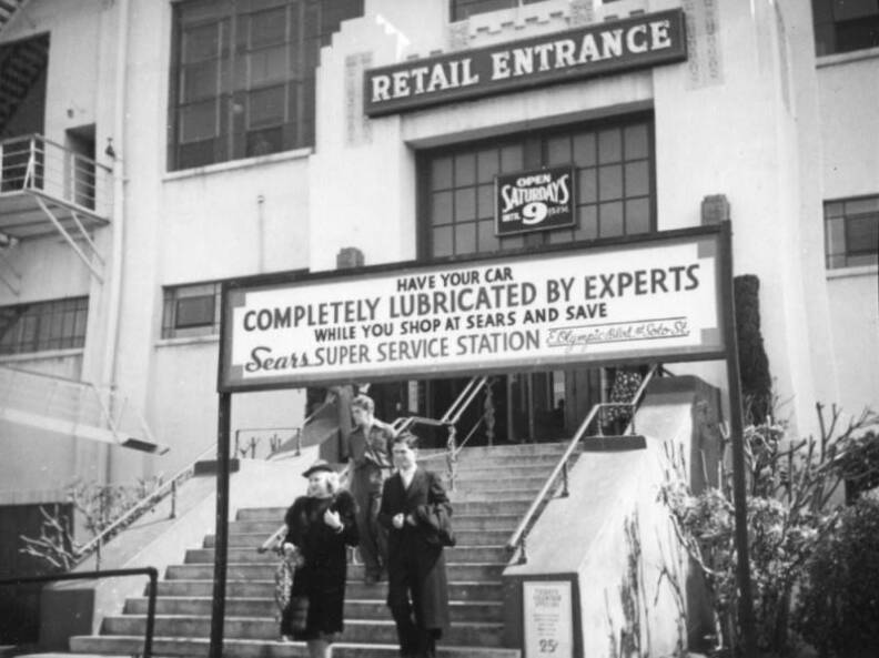 People walk down the stairs from the retail entrance to the Sears building in Boyle Heights circa 1938. Multiple signs indicate that the store was open past 9pm on Saturdays, that there was a 25-cent fountain special, and that you could get a lube job while you shop at their service station.