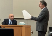 The setting is a courtroom: A man wearing a dark suit is sitting and looking at a man, also wearing a dark suit, as the man is speaking in reference to some papers in his hand. 