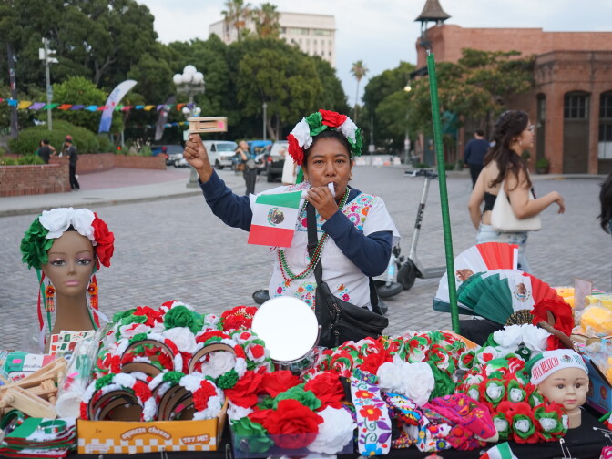 A woman with medium-dark skin and her hair up in a bun stands in front of a table full of Mexico merchandise. She holds a small Mexican flag.