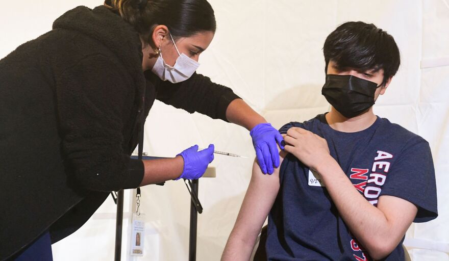 A medical practitioner wearing a mask and gloves gives an injection to a masked young man who is holding up his right sleeve.