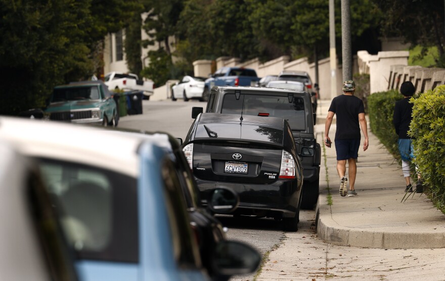 Two people are walking on a paved sidewalk in a residential neighborhood past a line of parked cars.