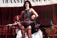 Tim Curry as Dr. Frank-N-Furter in The Rocky Horror Picture Show. He wears curly black hair, a white pearl necklace, a sparkly black vest, black panties, black gloves, and black thigh-high stockings.
