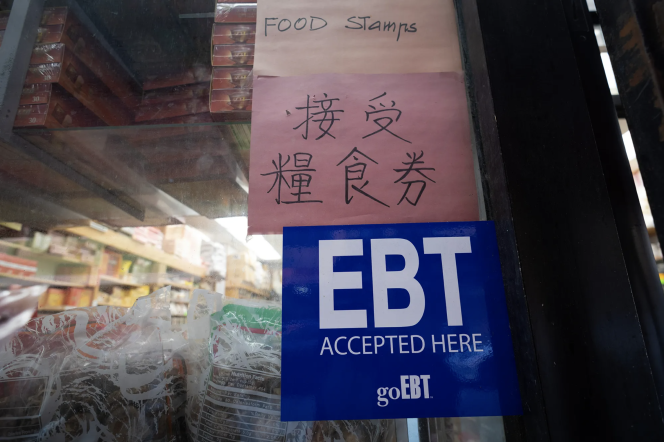 A sign that reads "EBT Accepted Here" hangs in the window of a shop. It hangs below two pink signs, one with Chinese characters and aonther with the words "Food Stamps." Plastic wrapped goods are lined up against the window.