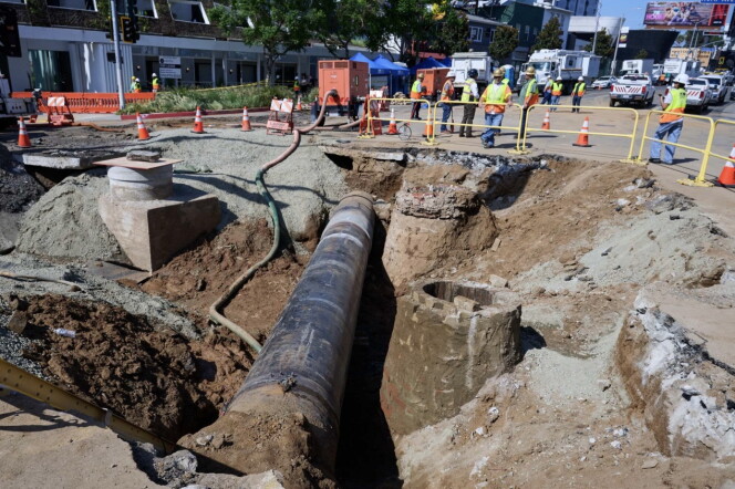 A section of a city street has been dug up, creating a deep trench with an exposed pipe running down the middle. 