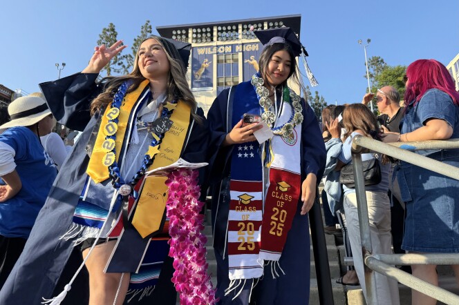 Two young women in graduation regalia walk through a crowd of people gathered in stands.
