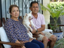 An older woman and man with light brown skin tone sit on chairs on a porch. The woman holds a white chihuahua in her left arm. 